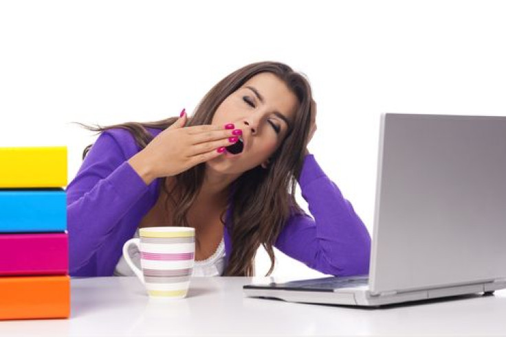 Woman yawning with coffee, books, and laptop on desk