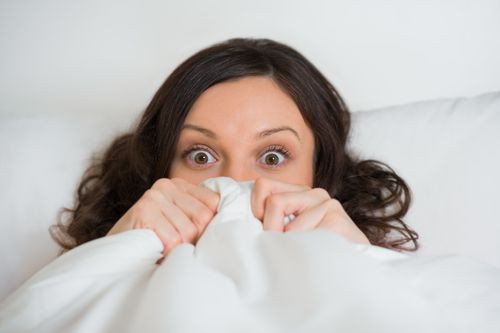 Woman hides her face in bed and looking at camera with wide eyes