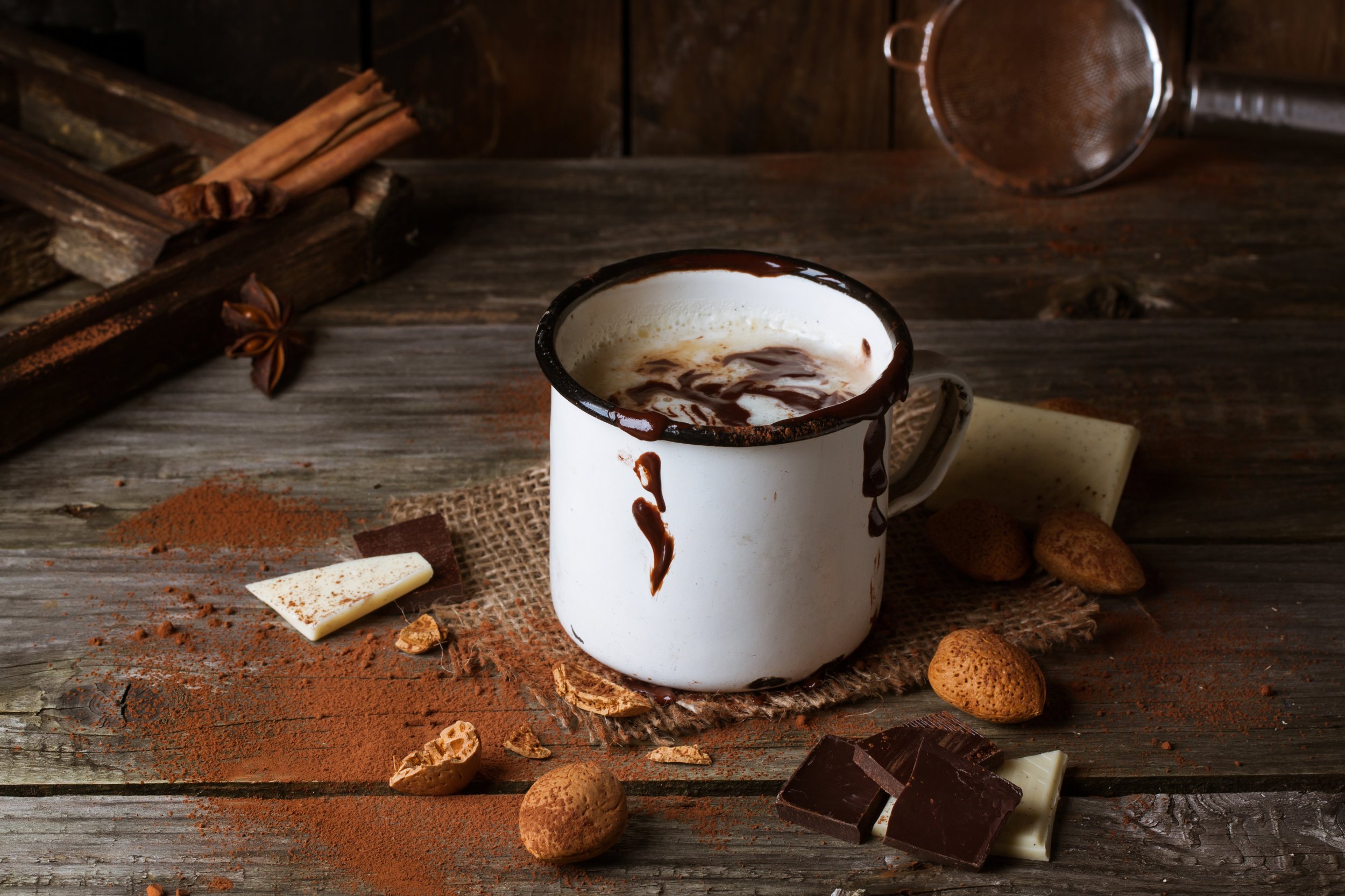 Mug with hot chocolate served with chunks of white and dark chocolate and almonds on old wooden table
